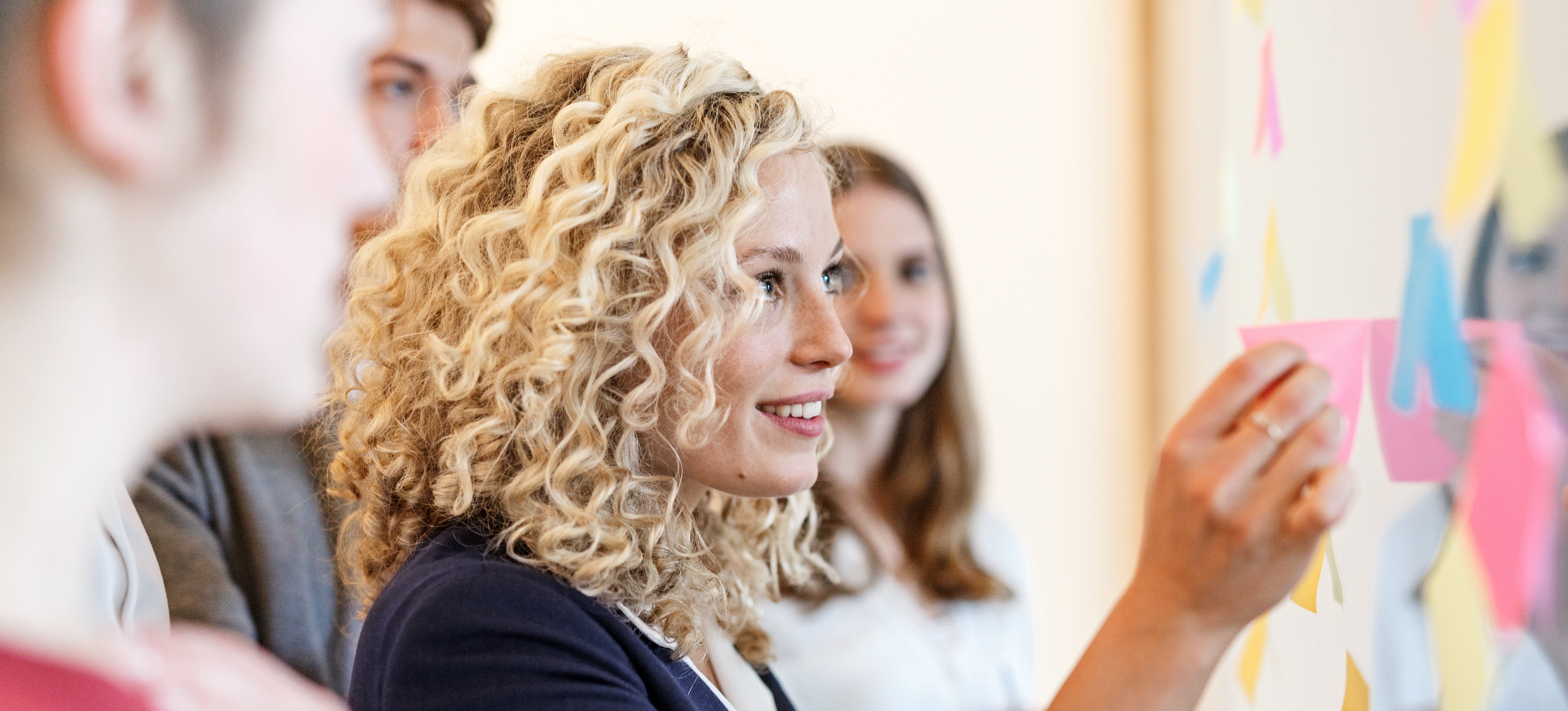 [Featured Image] A content strategist talking to three colleagues moves a sticky note on a board covered with many other, multi-colored sticky notes.
