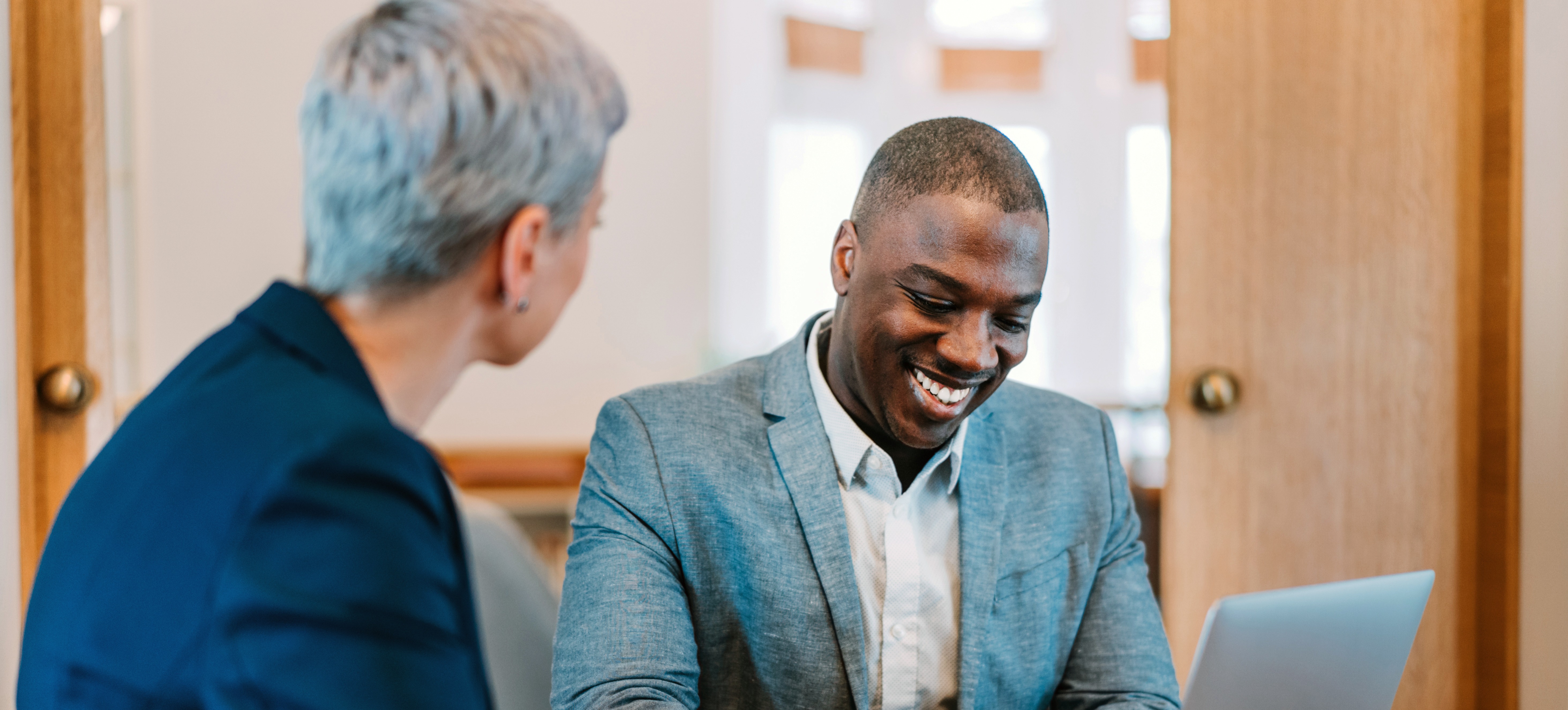 [Featured Image] A person holding an entry-level analyst job, wearing a gray sports jacket and white shirt, is working on their laptop and conferring with a co-worker in their office.