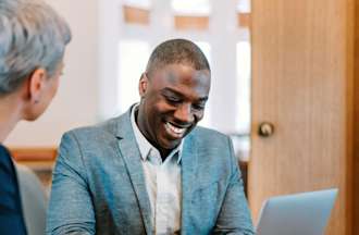 [Featured Image] A male, wearing a gray sports jacket and white shirt, is working on his laptop and conferring with a co-worker in his office.