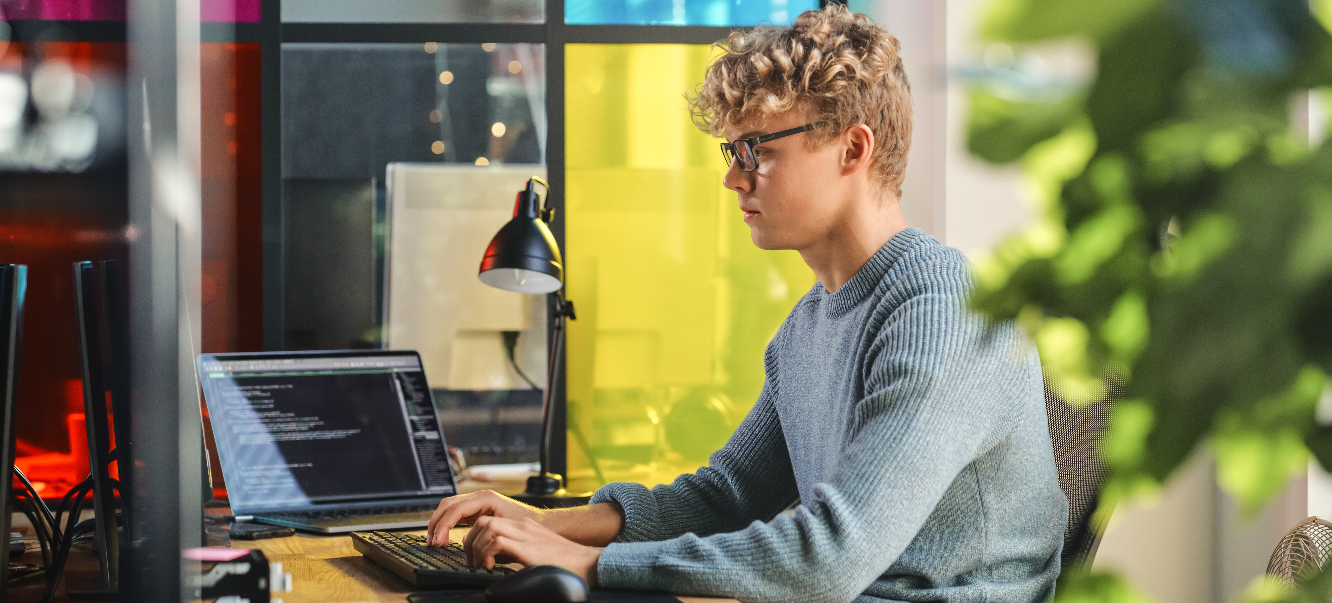 [Hauptbild] Ein junger Programmierer verwendet die Programmiersprache Julia, während er an zwei Computern in einer farbenfrohen, ungezwungenen Büroumgebung an Code arbeitet.