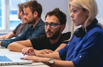 [Featured image] Master's degree students study in a computer lab, sitting along a long table in front of laptops. One student kneels alongside another and they share a screen.