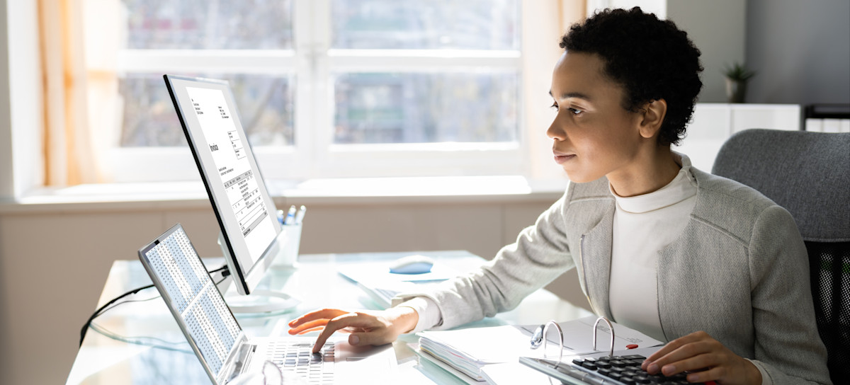[Featured Image] A woman who receives an accountant salary works on her laptop and calculator simultaneously at her desk.
