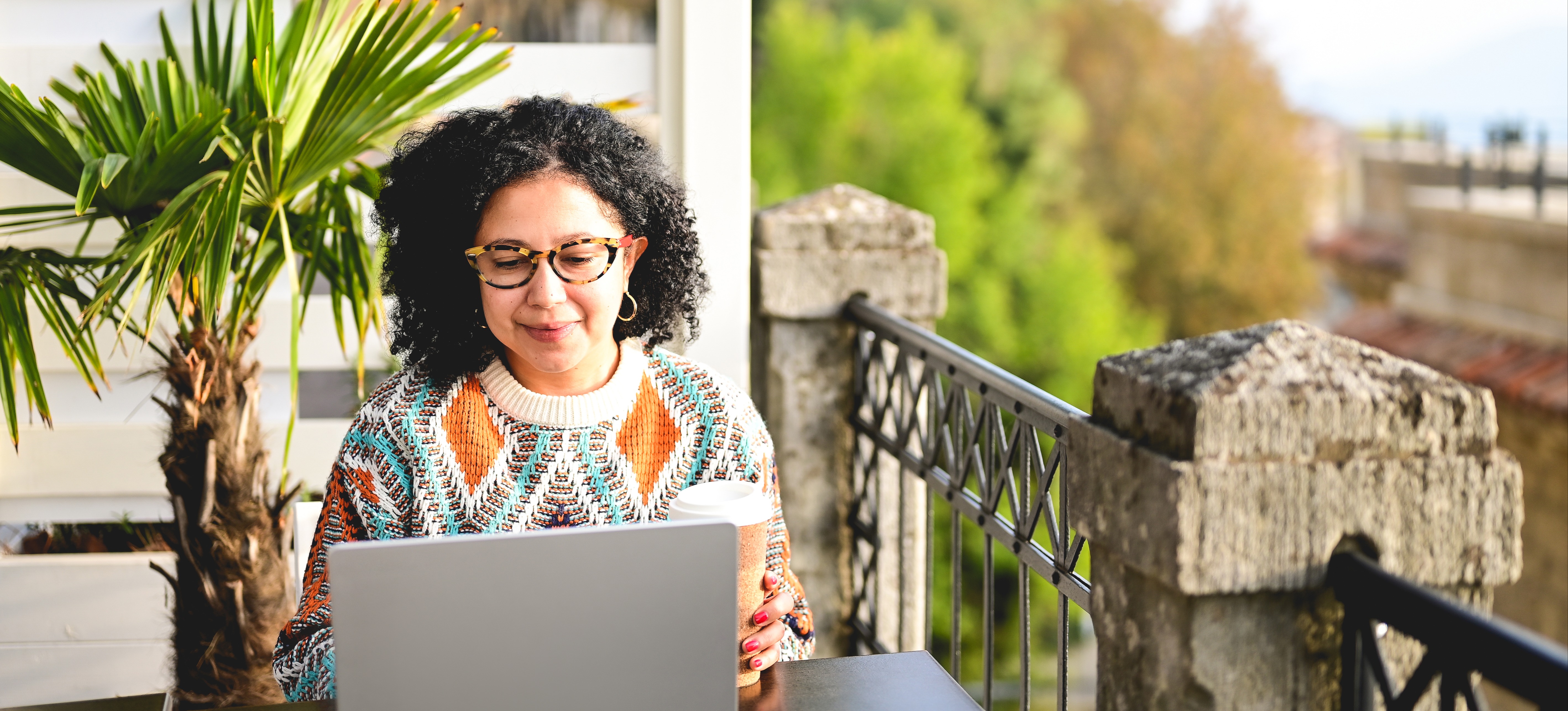 [Featured Image] A women sits at a table on her balcony while looking at her laptop. She is researching the difference between chatbot vs conversational ai.