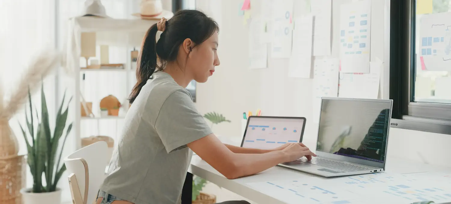 [Featured Image] A person sits in their sunny home office and uses sparklines in Google Sheets on their laptop and a second device.