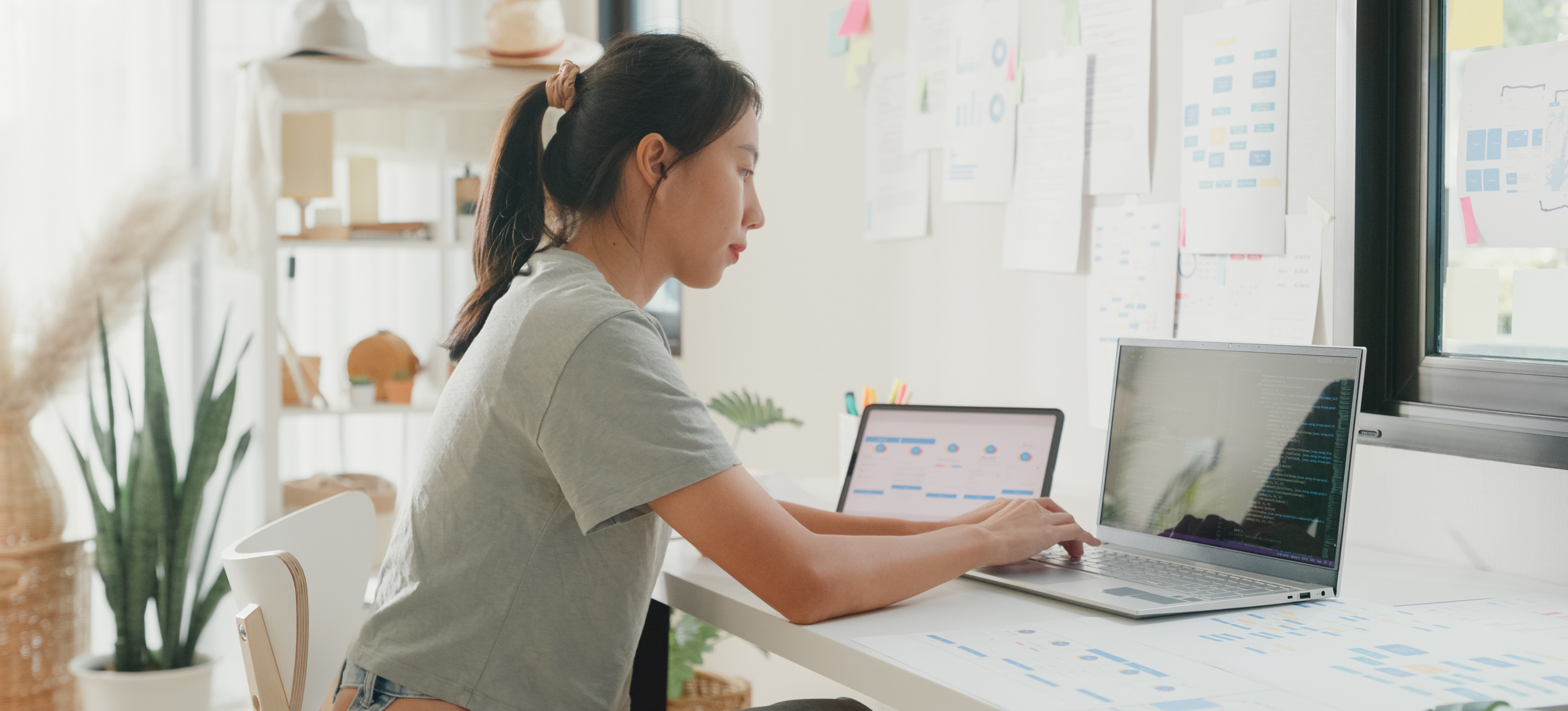 [Featured Image] A person sits in their sunny home office and uses sparklines in Google Sheets on their laptop and a second device.