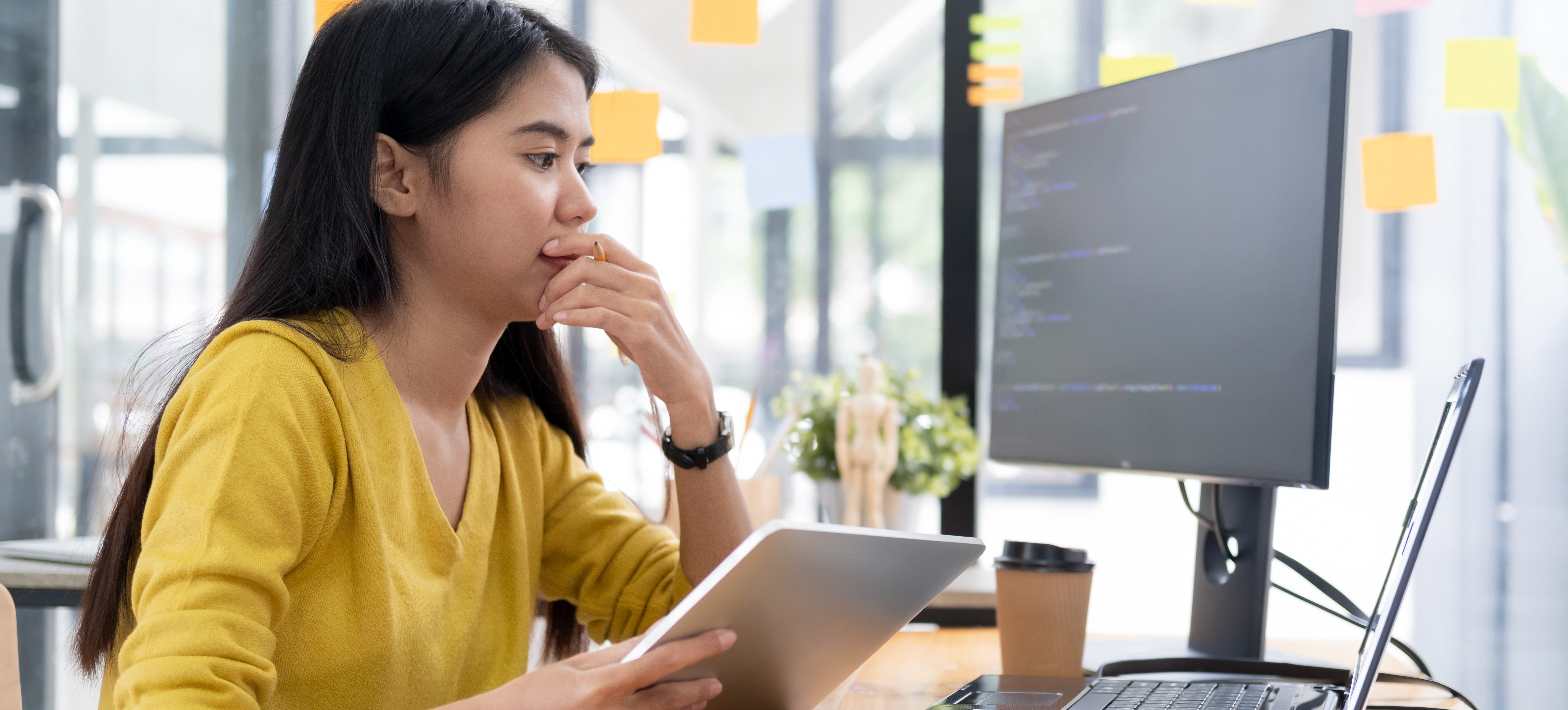 [Featured Image] A young programmer who discovered how to learn SQL sits at her office desk with a laptop and tablet and works.
