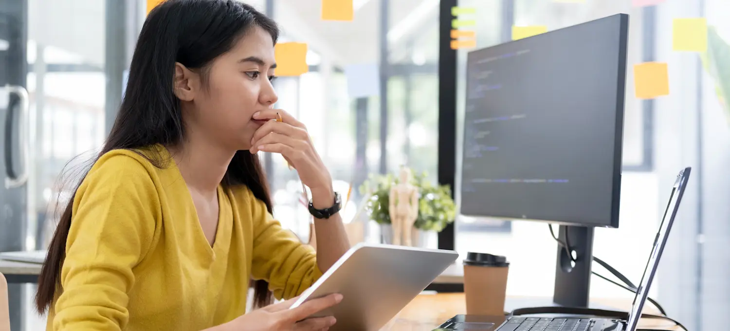 [Featured Image] A young programmer who discovered how to learn SQL sits at her office desk with a laptop and tablet and works.
