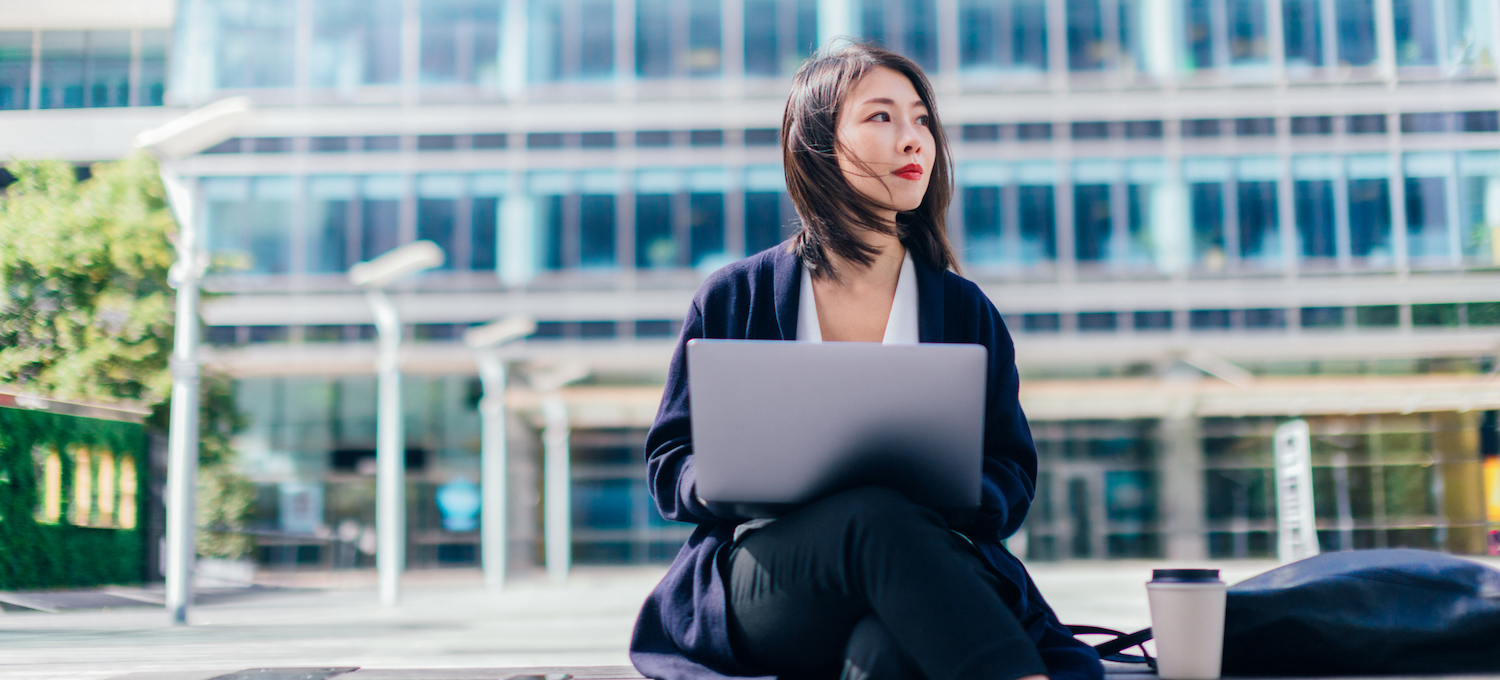 [Featured image] A young professional wearing a black trench coat sits outside using their laptop.