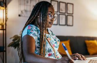 [Featured image] A Black woman in glasses sits at home taking notes off something she reads on her laptop.
