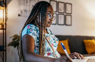 [Featured image] A Black woman in glasses sits at home taking notes off something she reads on her laptop.