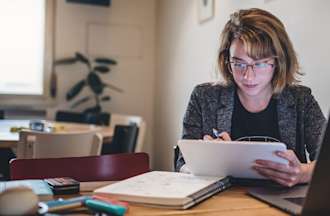 [Featured image] A woman sitting in front of a laptop and a notebook uses her tablet. 