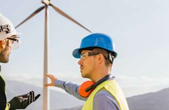 [Featured Image] A renewable energy engineer talks to a technician at a wind turbine.  