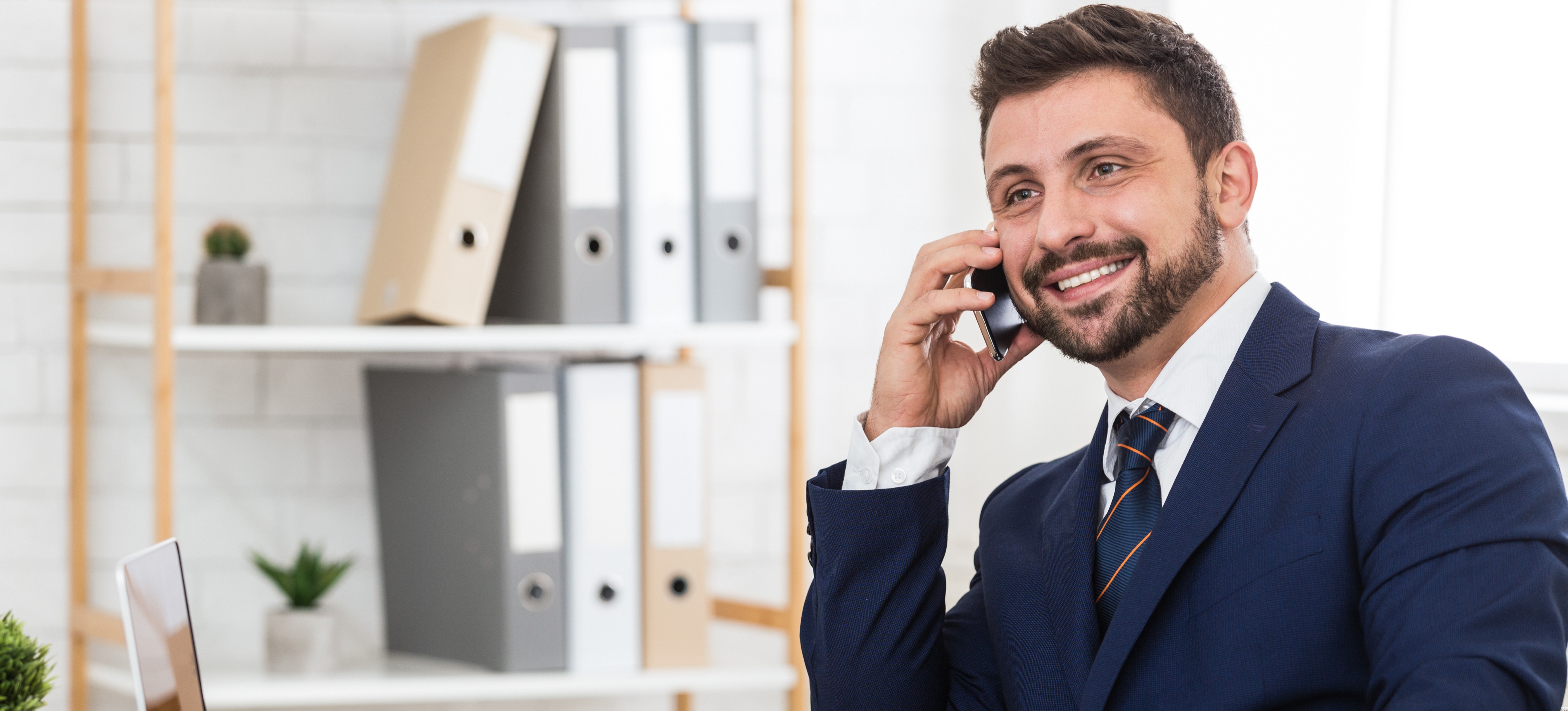 [Featured image] A professional-looking business person who has earned a recruiter degree and is dressed in a blue suit and tie smiles as they talk on the phone with a potential employee while sitting in their office.

