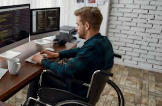 [Featured Image] A man using a wheelchair works at a desktop computer with two monitors. An open laptop rests on the left of the desk. 