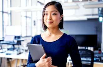 [Featured image] A cybersecurity engineer in a blue long-sleeve shirt stands in an open office space holding their tablet.