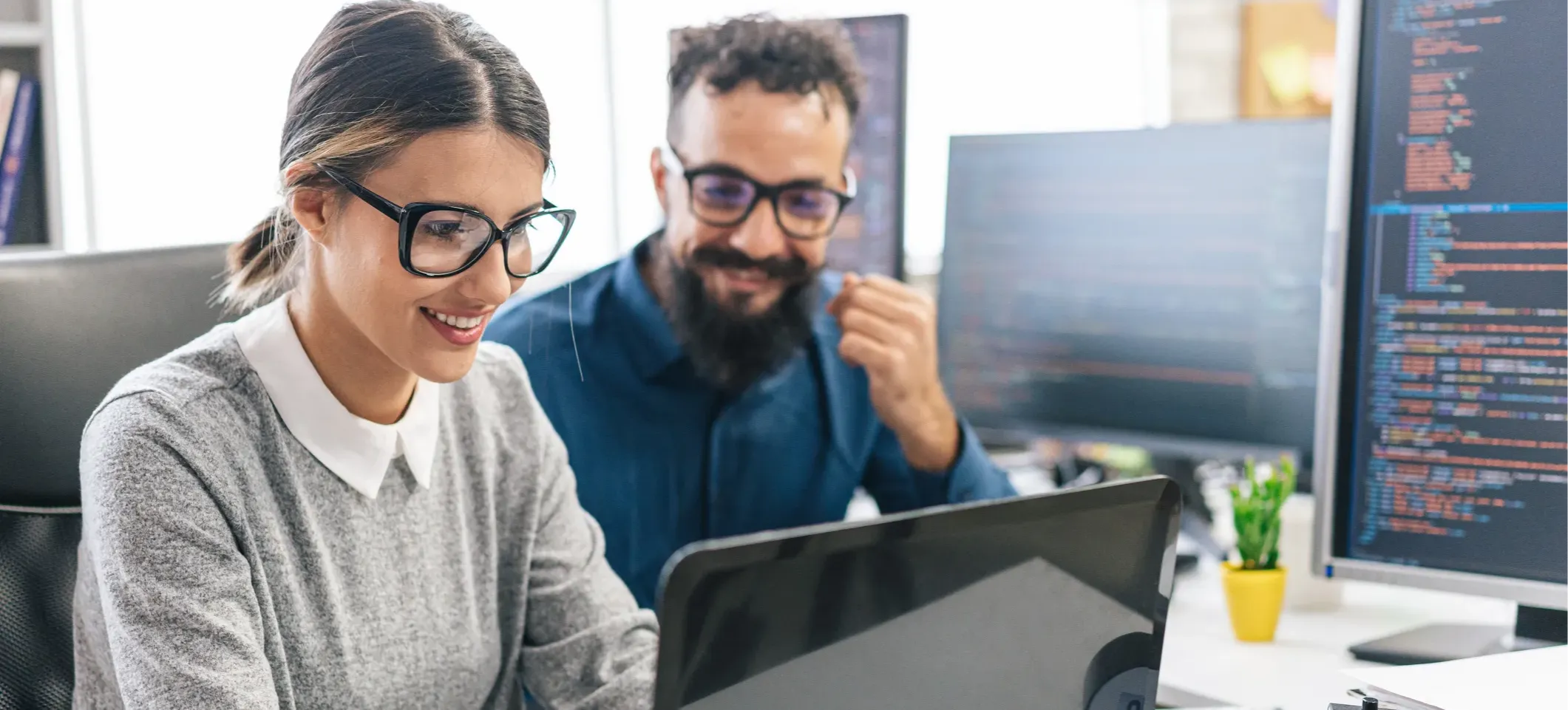 [Featured Image] Two coworkers are sitting at a desk, looking at a laptop and discussing possible cybersecurity career paths. Three additional computer monitors with code appear in the background.
