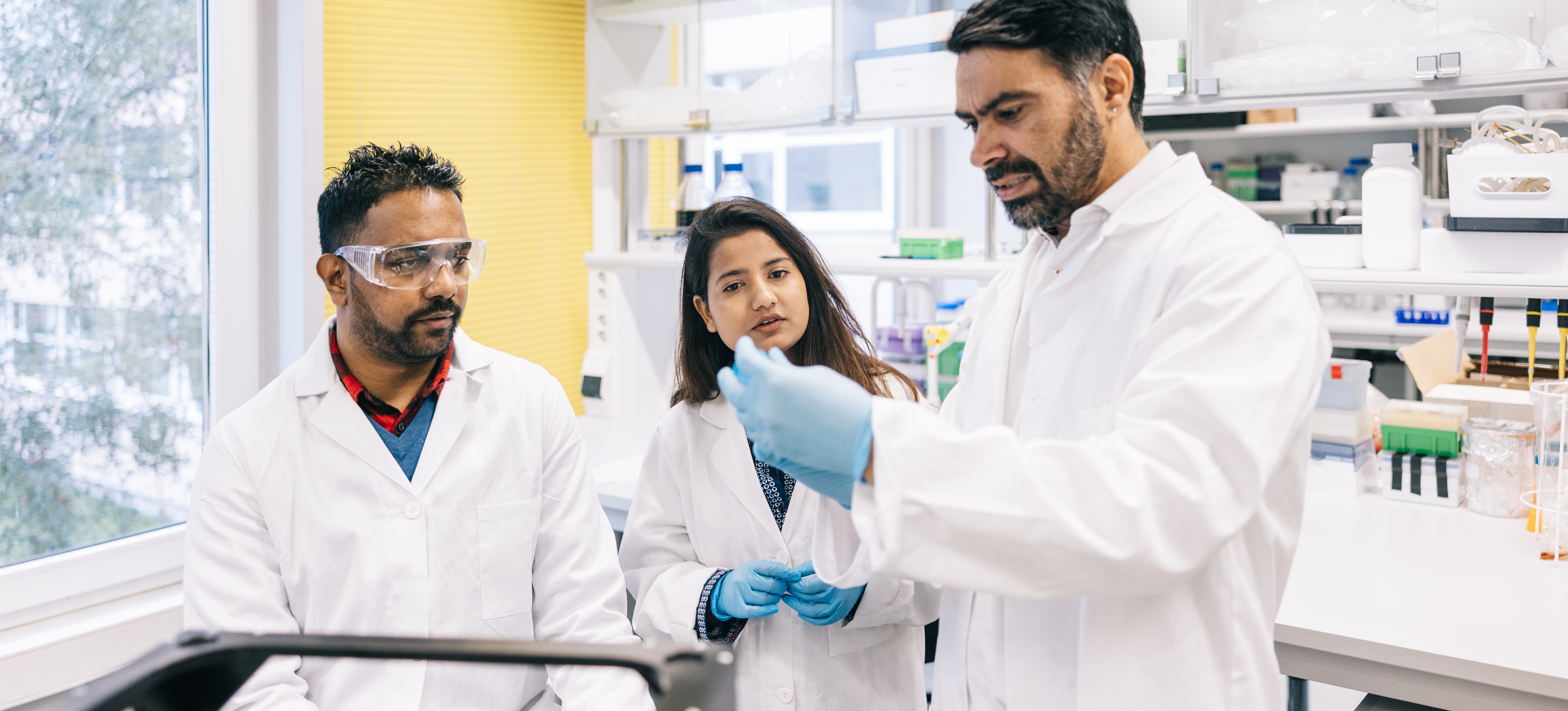 [Featured Image] A clinical research associate stands in a lab with two medical professionals who are discussing clinical trials.
