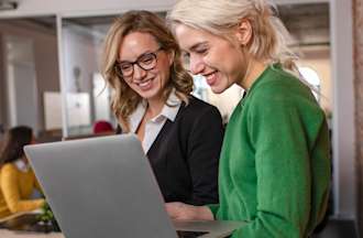 [featured image] Two blond women stand looking at a laptop screen.