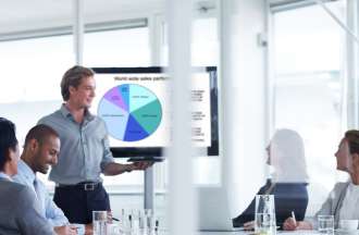 [Featured image] A male product promoter, wearing a blue shirt and standing in front of a  screen that shows a pie chart.  He is conducting a meeting, as he promotes the latest product being offered.  
