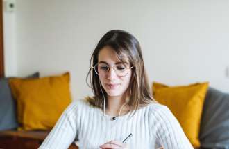 [Featured Image]:  A female wearing a white top and glasses. She has long brown hair. She is sitting in front of her laptop, holding a pen in her hand. 