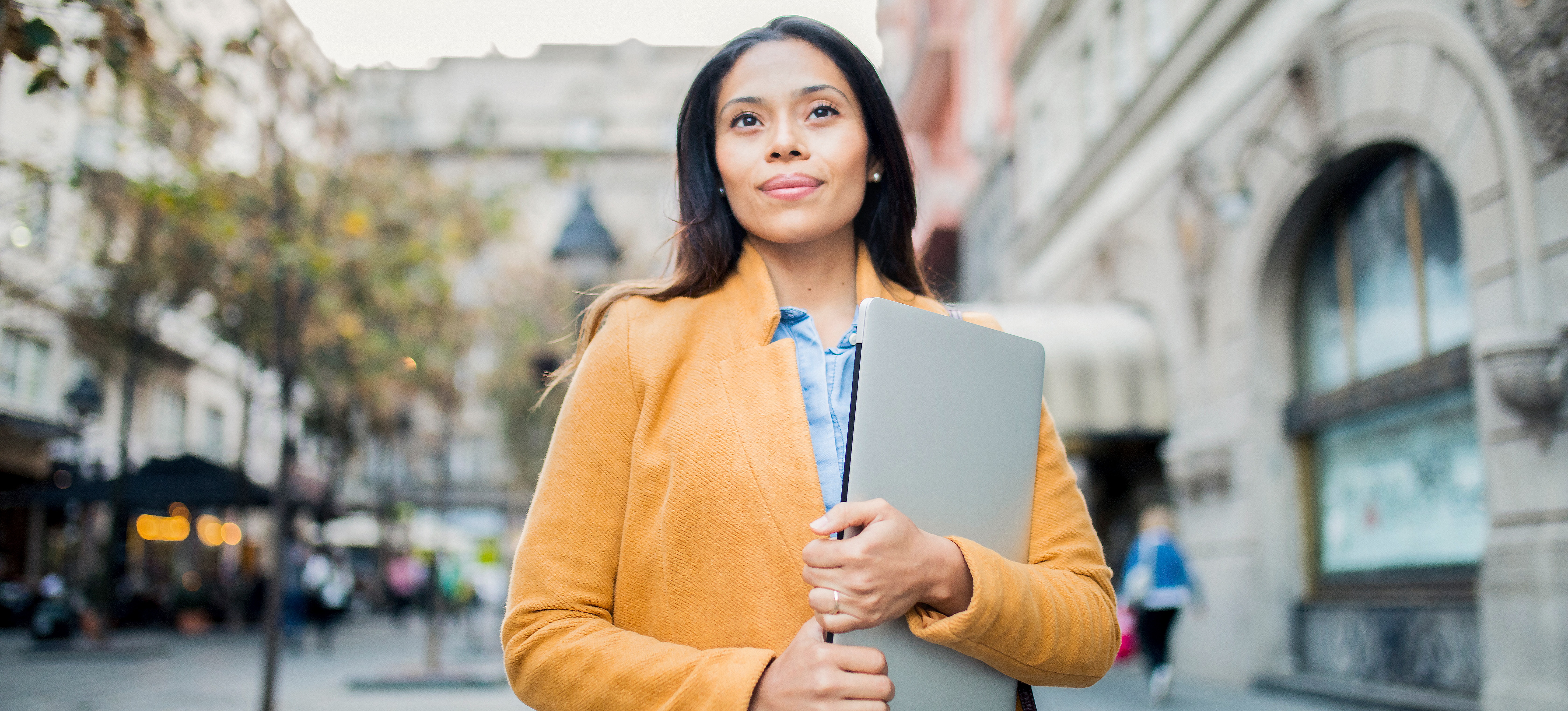 [Featured Image] A person stands outside an office building holding a laptop computer.