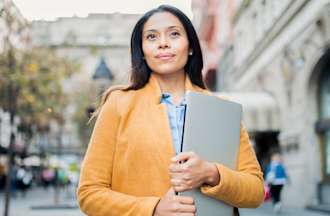 [Featured Image] A woman stands outside an office building holding a laptop computer.