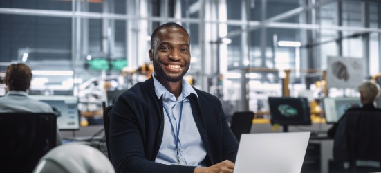 [Featured Image]:  A male data engineer is wearing a blue shirt and dark jacket.  He is working  at his laptop, at his desk in an office with other team members. 
