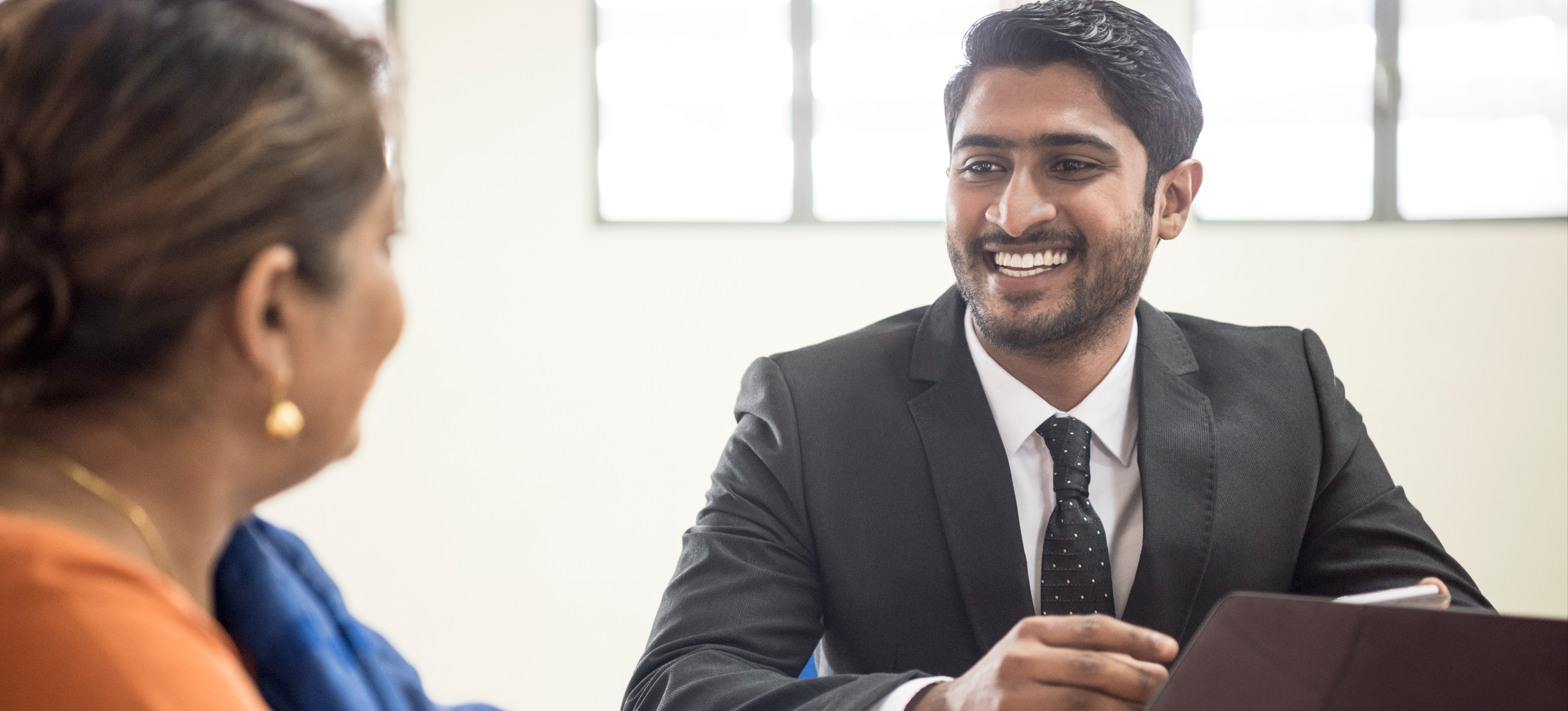 [Featured Image] A smiling IT business analyst talks to a client across a table as they are both using laptops.

