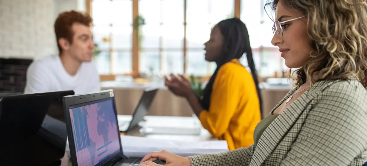 [Featured Image] A businessperson working for a network analyst salary sits at a coworking table beside two colleagues and types on their laptop.
