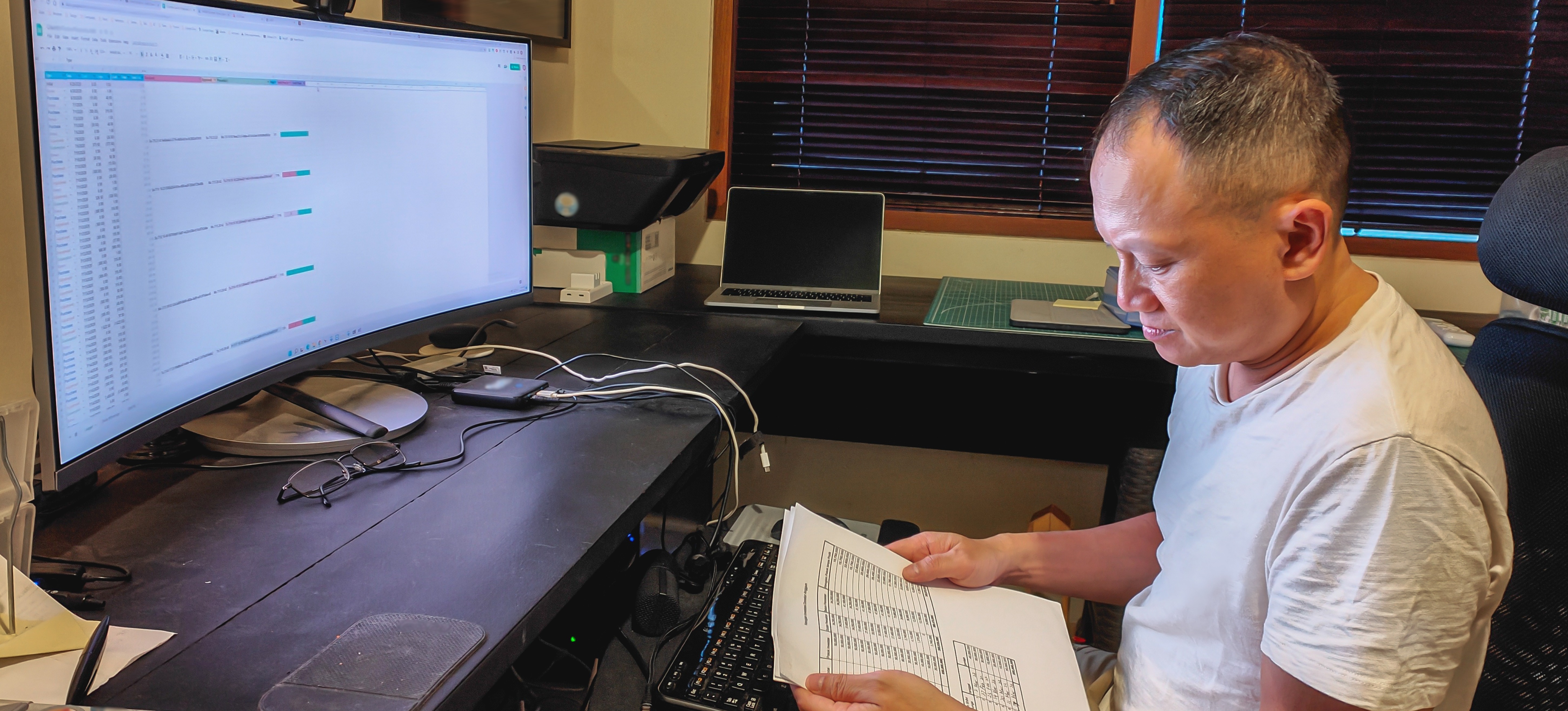 [Featured Image] A person holds a printout of an Excel spreadsheet while sitting at their desk, with the spreadsheet displayed on their computer monitor.