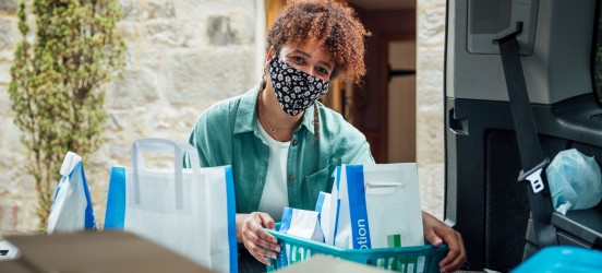 [Featured Image] A community health worker in a green jacket, white top, and a face covering loads prescription medication into a van.