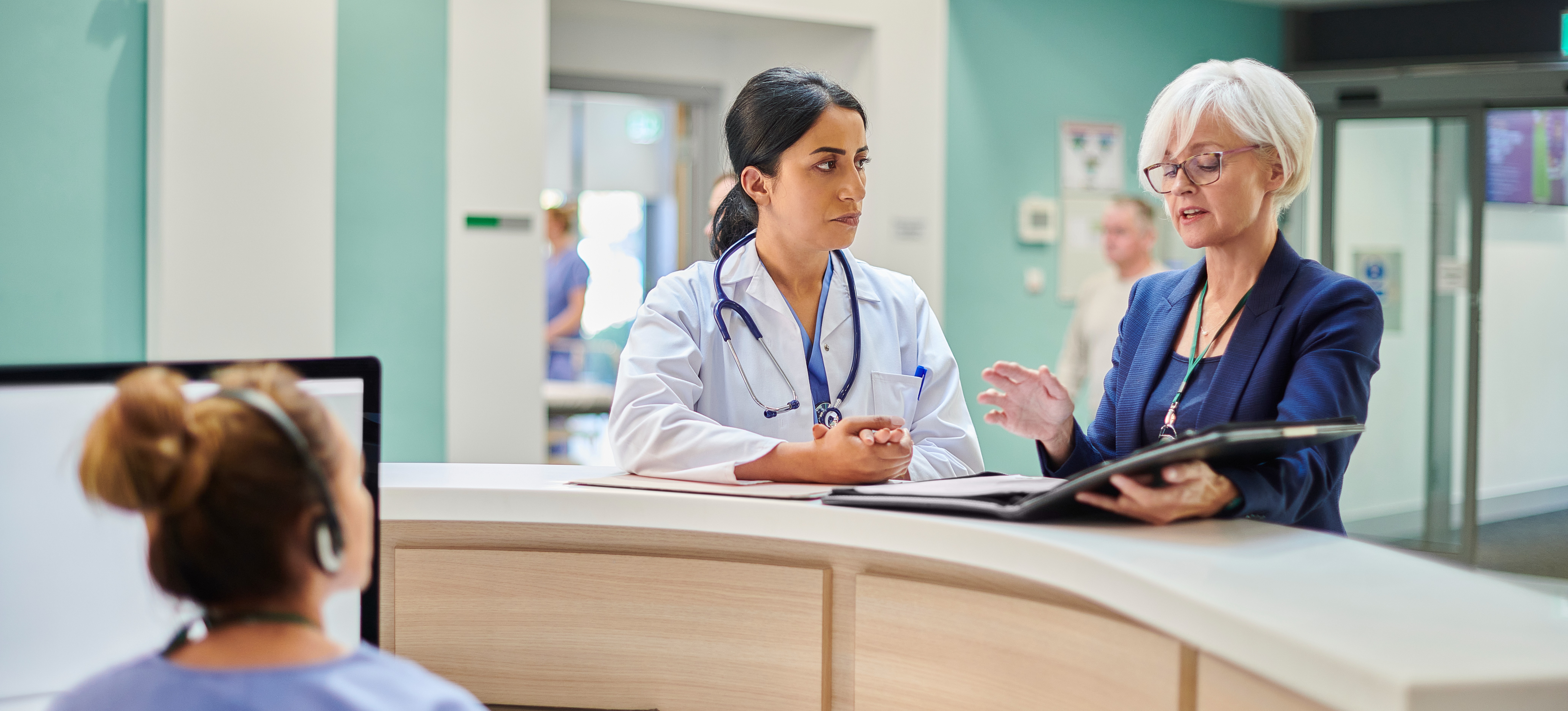 [Featured Image] A health care administrator holds a binder of notes while working alongside a doctor at a hospital.
