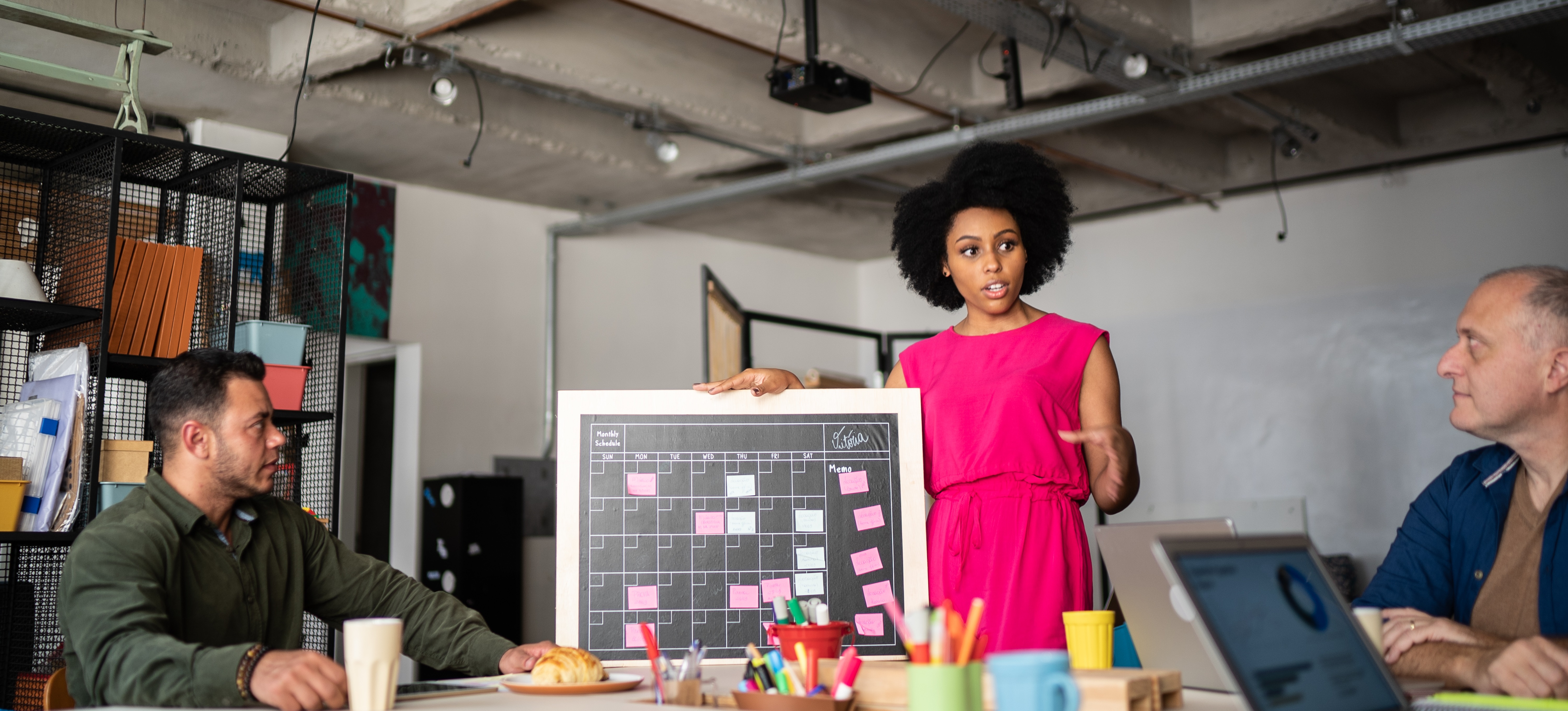 [Featured Image] A person working for a marketing manager addresses two of their colleagues at a meeting table as they hold a calendar and discuss their upcoming marketing plan.
