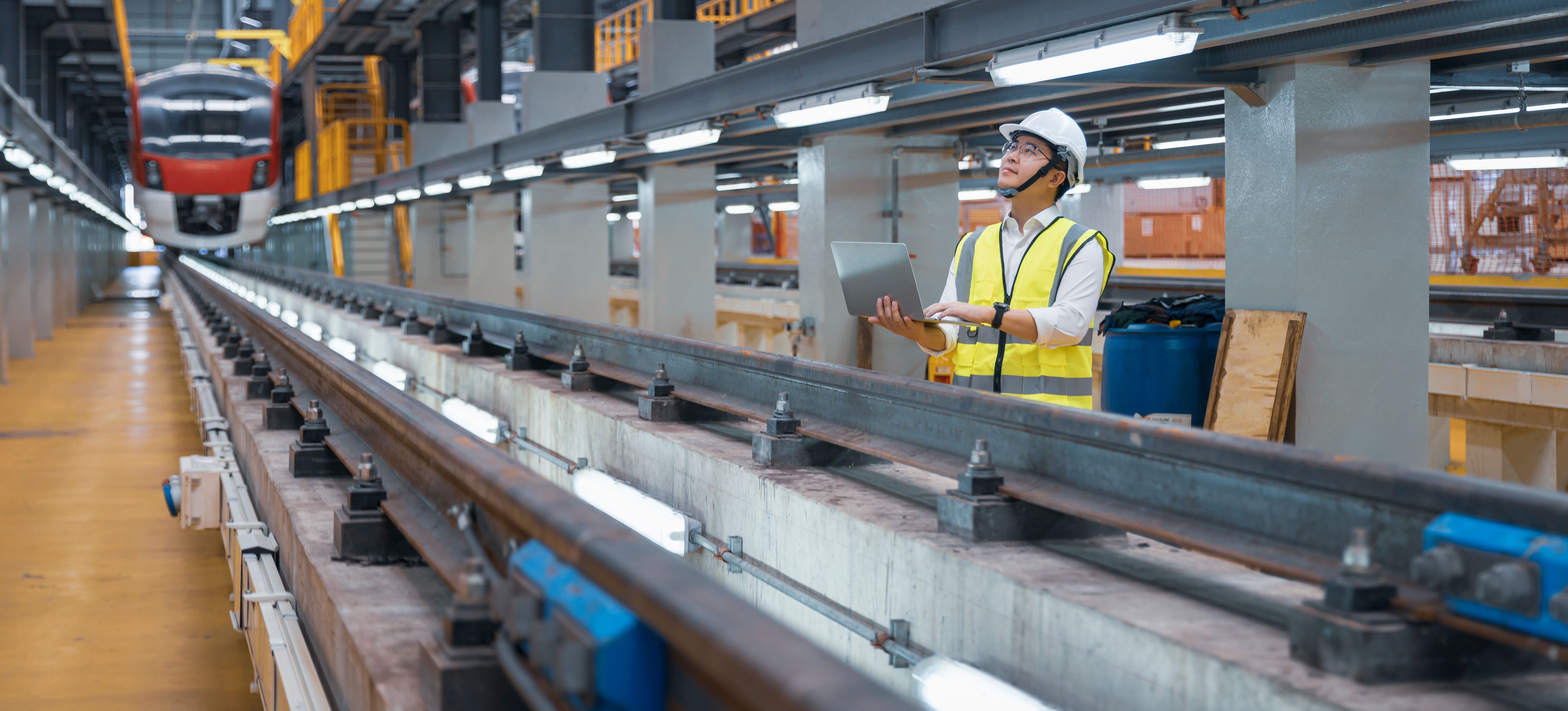 [Featured Image] An operations engineer in the transportation industry holds a laptop while standing in a railway workshop.
