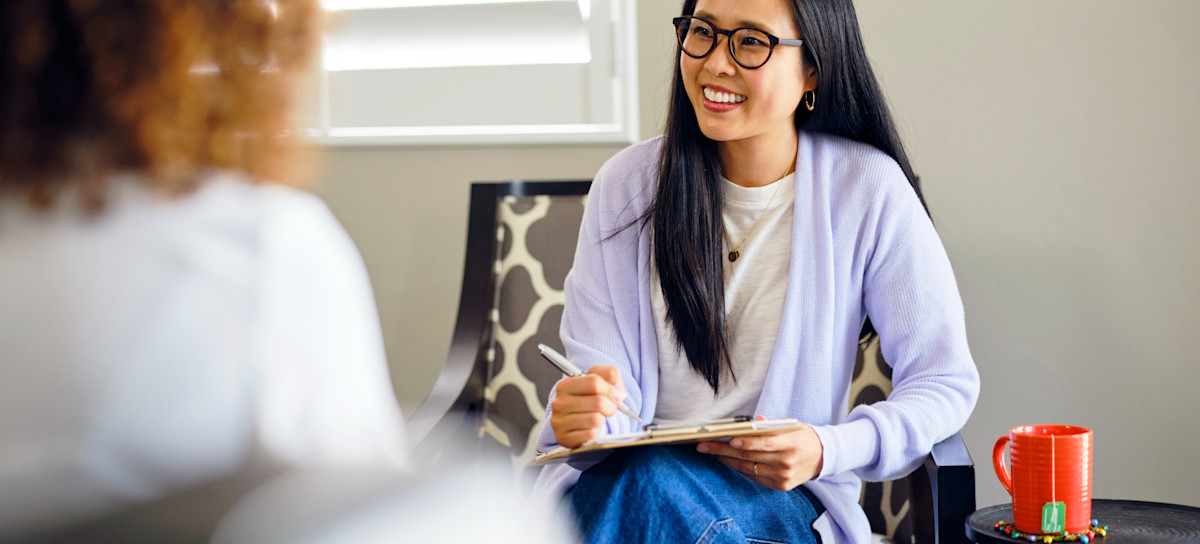 [Featured image] A certified life coach sits on a chair and holds a clipboard while speaking with a client. 
