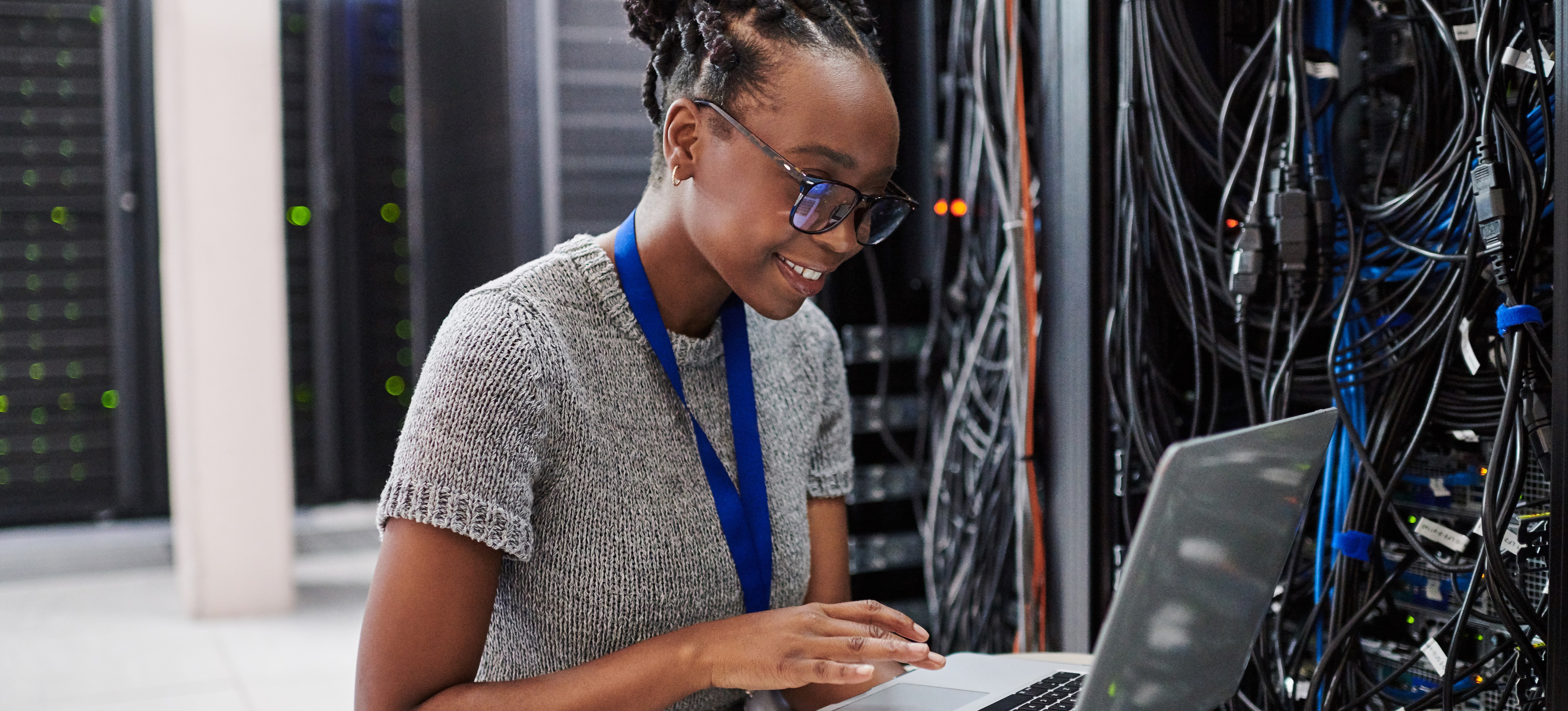 [Featured Image] Un ingénieur réseau souriant est assis sur le sol d'une salle de serveurs et travaille sur son ordinateur portable.