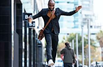 A smiling professional wearing a shoulder bag and carrying a to-go cup of coffee leaps from one bench to the next outside an office building.