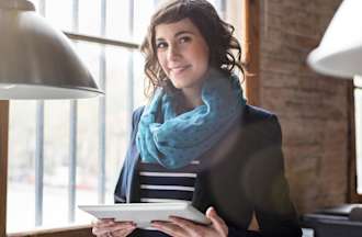 [Featured image] A woman working as a social media manager stands by a window with her tablet.