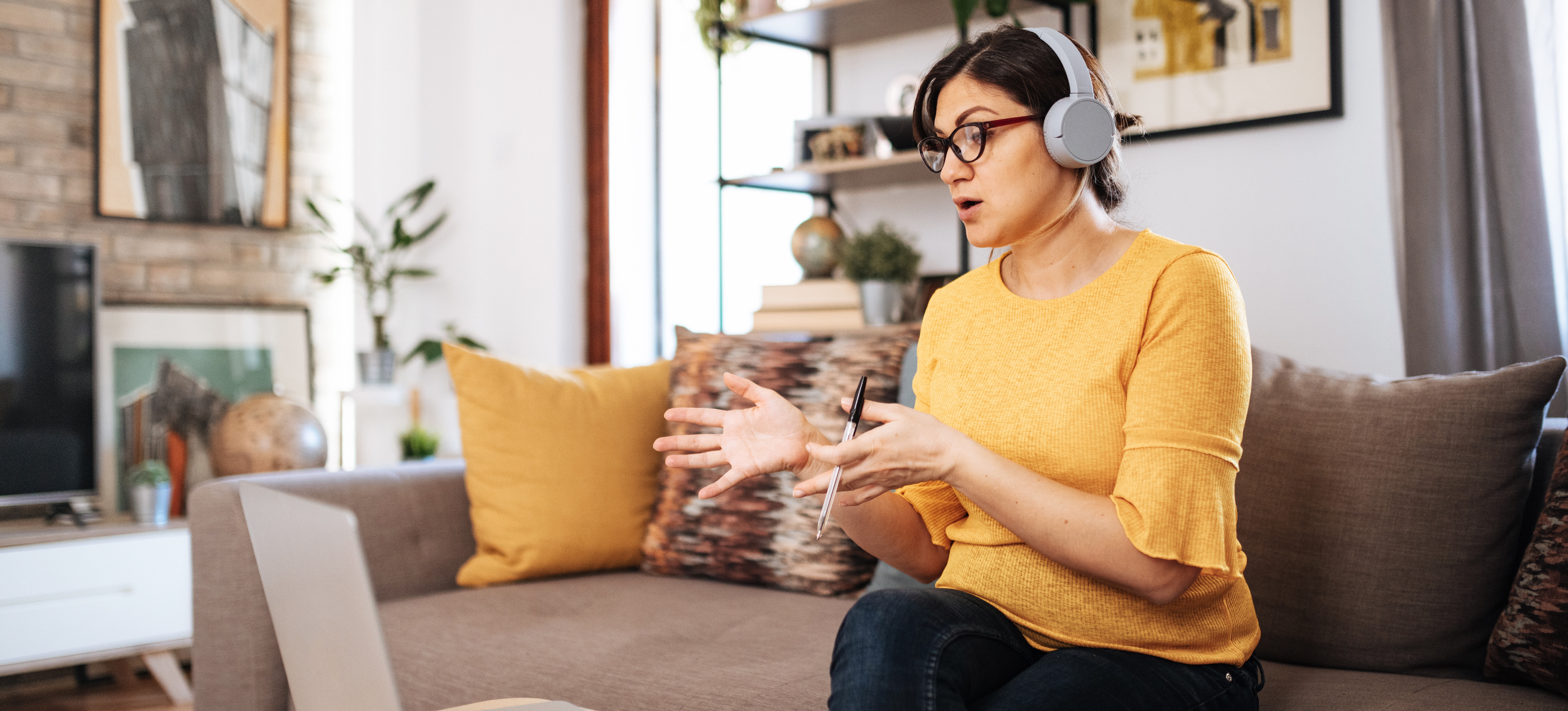 [Featured Image] Social media manager teleconferencing with a client while sitting on a sofa with their laptop and an open notebook on the table in front of them. 
