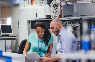 [Featured Image]:  A male, wearing a blue shirt and a female, wearing a green uniform, are discussing records and charts in a room  with computers and big data information.