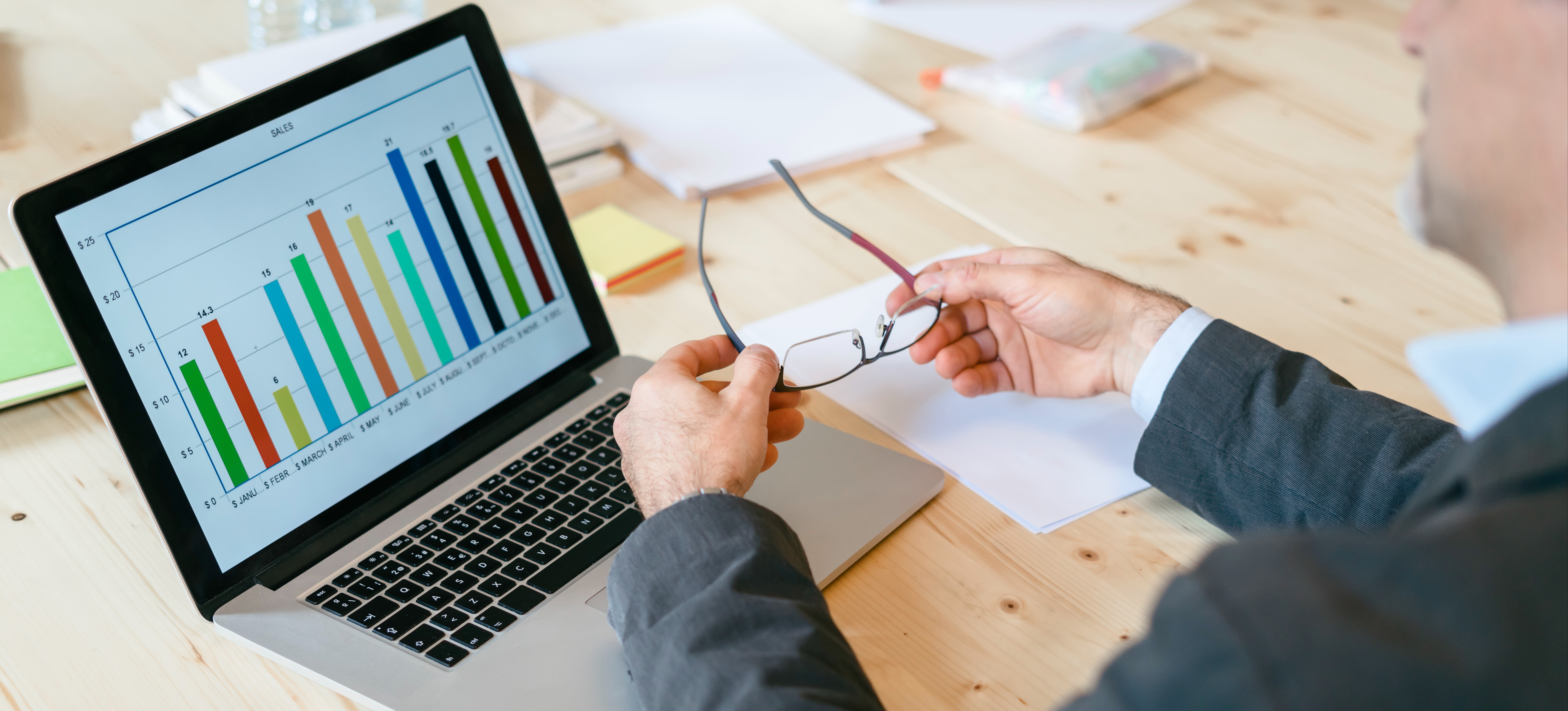 [Featured Image] A business professional removes their glasses to examine a bar chart on a laptop.  