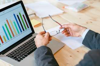 [Featured Image] A businessman takes off his glasses and examines a bar chart on a laptop.  