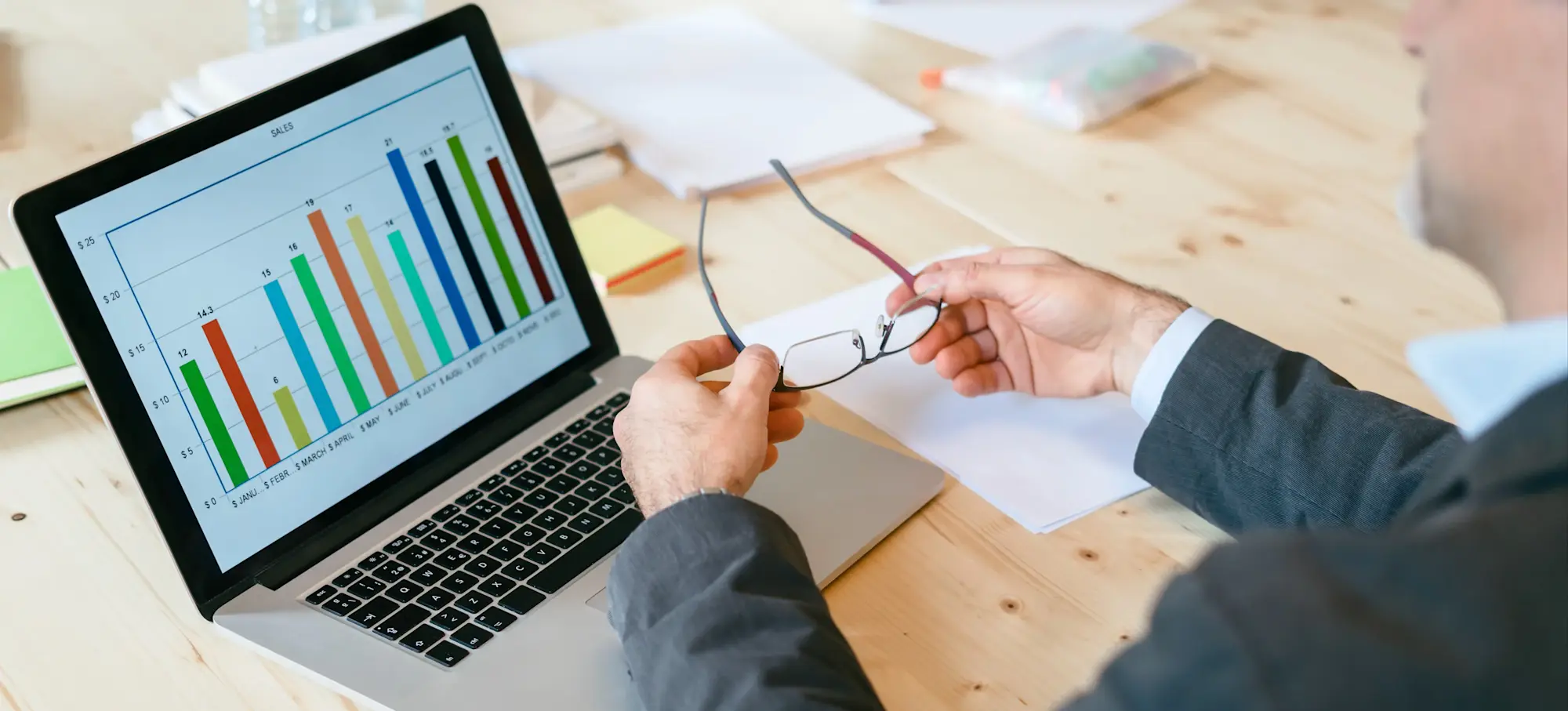[Featured Image] A business professional removes their glasses to examine a bar chart on a laptop.  