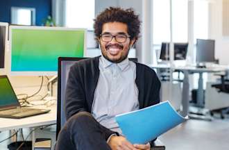 [Featured image] A man with glasses and a blue folder sits at a cybersecurity workstation with three computer screens.