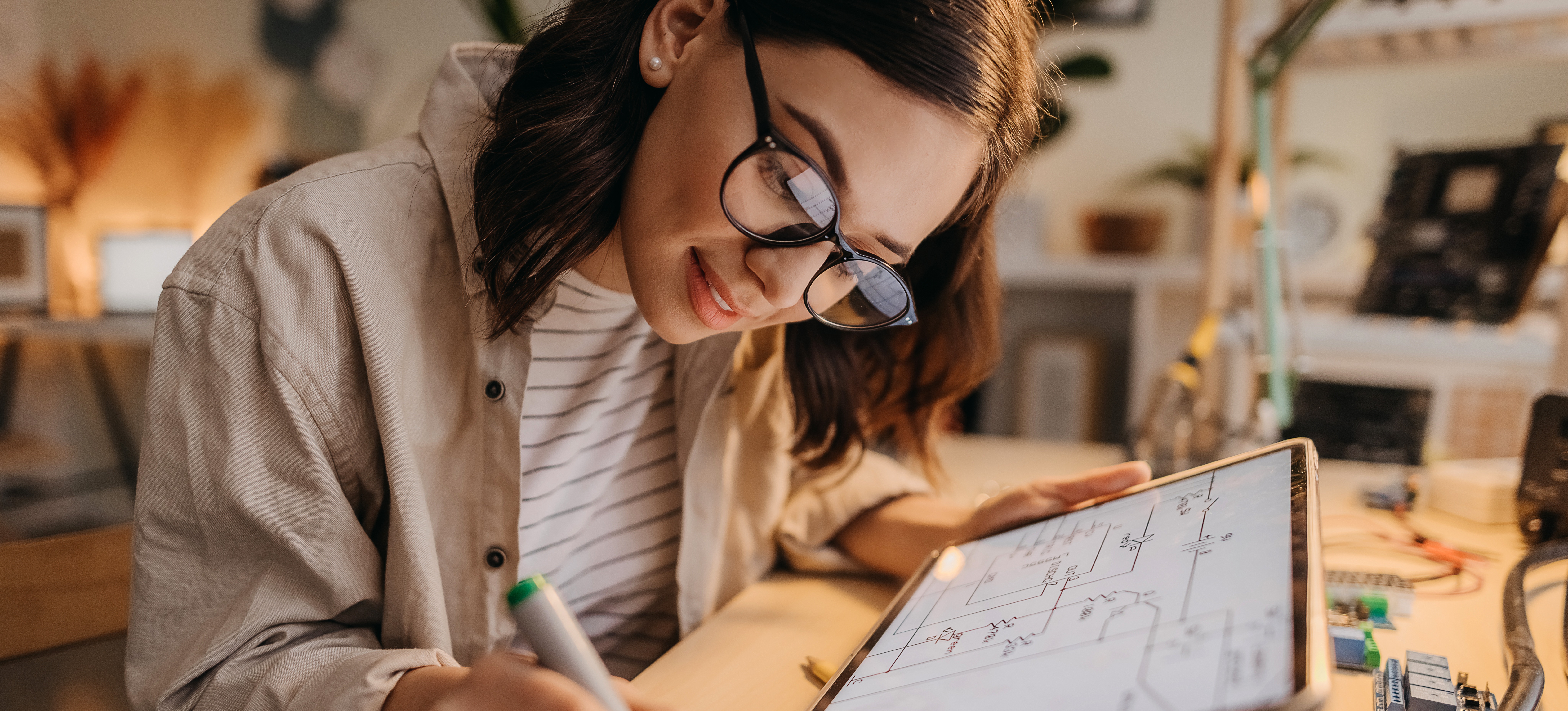 [Featured Image] A smiling person specializing in hardware engineering hovers over their desk, filled with hardware, as they hold a tablet and take notes.

