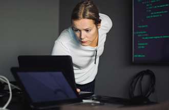 [Featured image] A cybersecurity professional in a white shirt stands in front of a laptop and wall monitor.