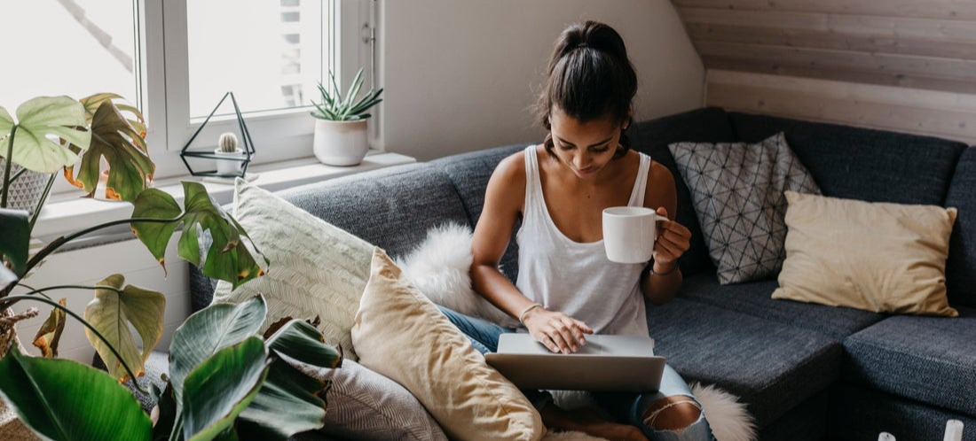[Featured image] A person sits on their living room couch next to a window with a coffee mug in hand as they work on their Shopify Etsy Integration on their laptop.