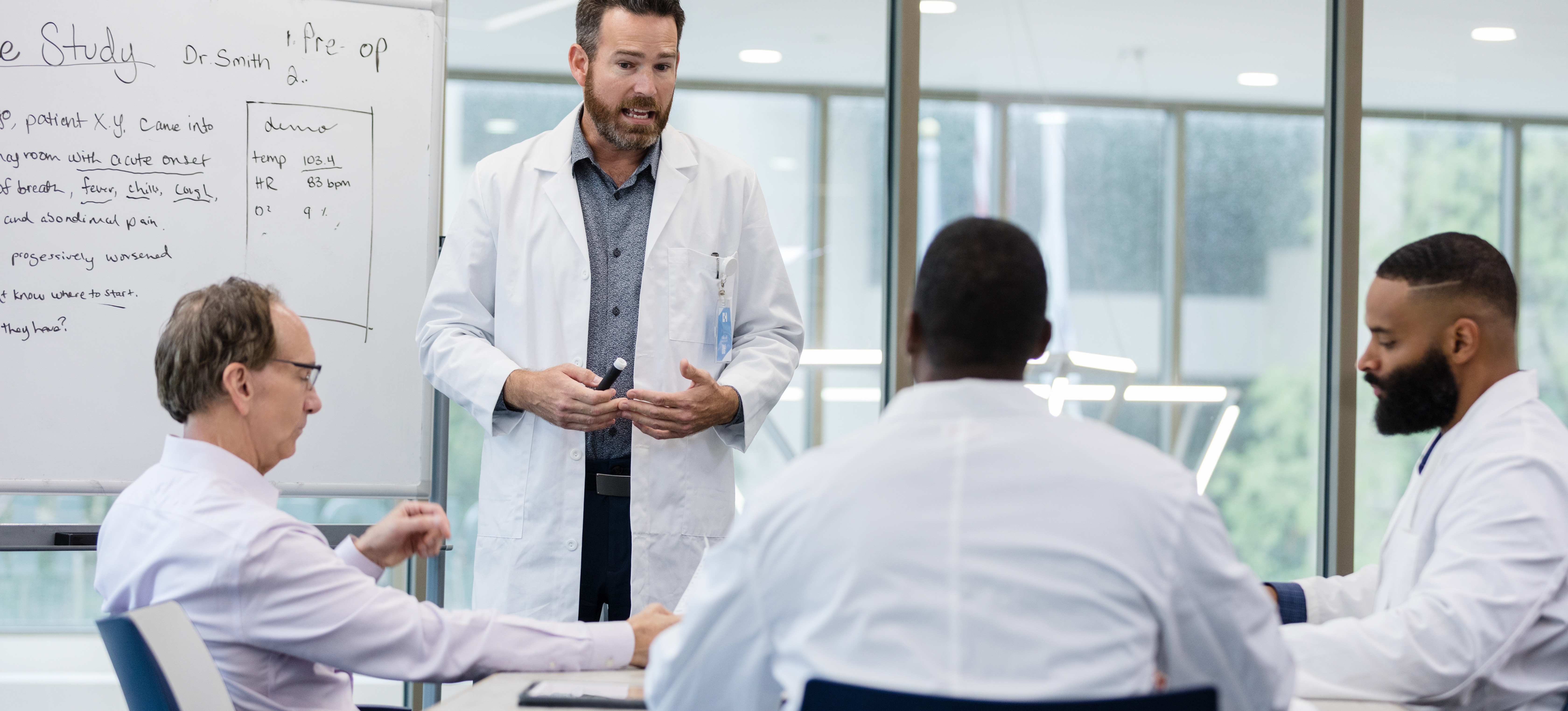 [Featured image] A public health analyst in a white lab coat talks with three hospital administrators in a windowed boardroom about a case study.