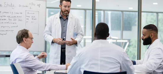 [Featured image] A public health analyst in a white lab coat talks with three hospital administrators in a windowed boardroom about a case study.