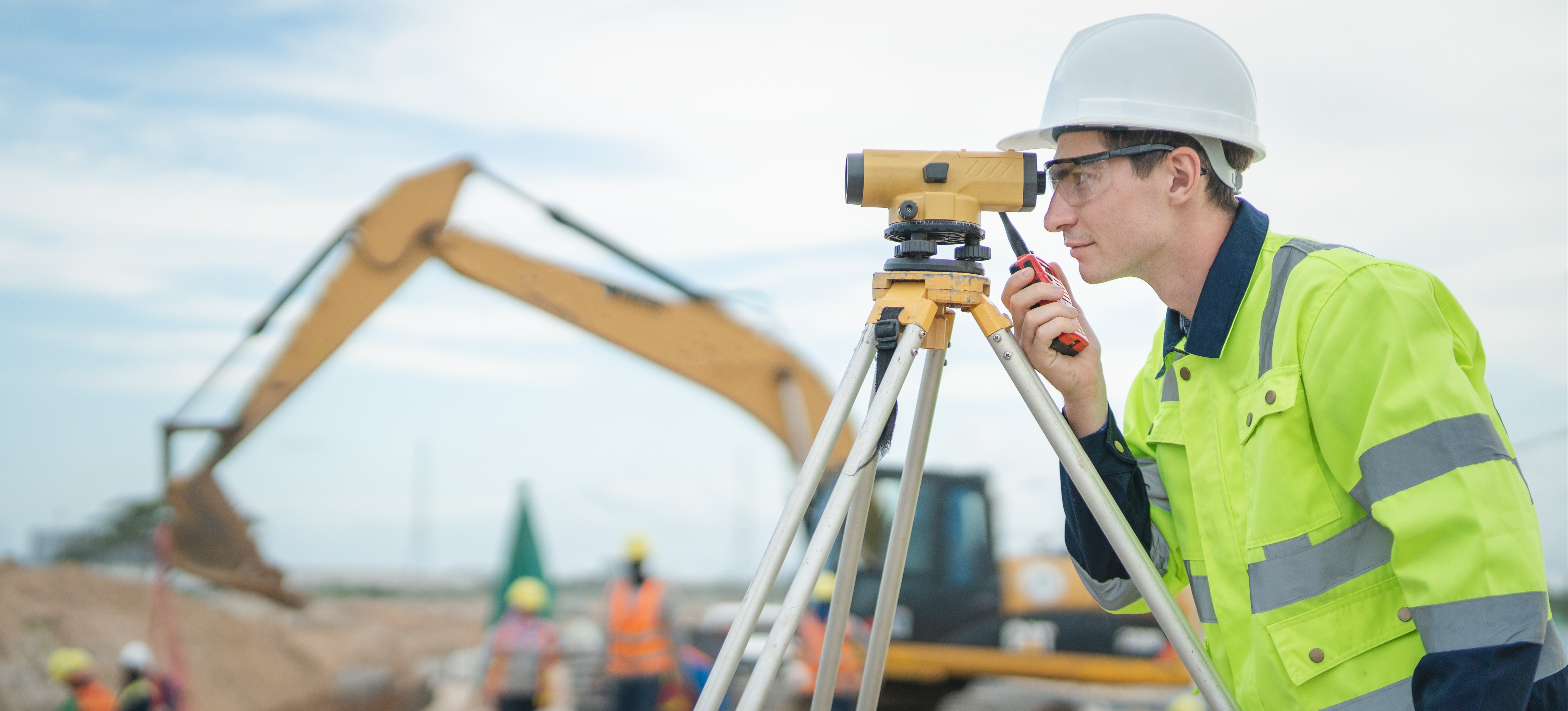 [Featured Image] An engineer wearing a hard hat and safety vest looks through survey equipment on a construction site.
